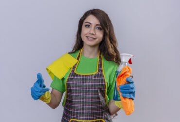 young beautiful woman with long wavy hair wearing apron and rubber gloves holding rug and cleaning spray looking confident and positive smiling showing thumbs up ready to clean standing over blue b